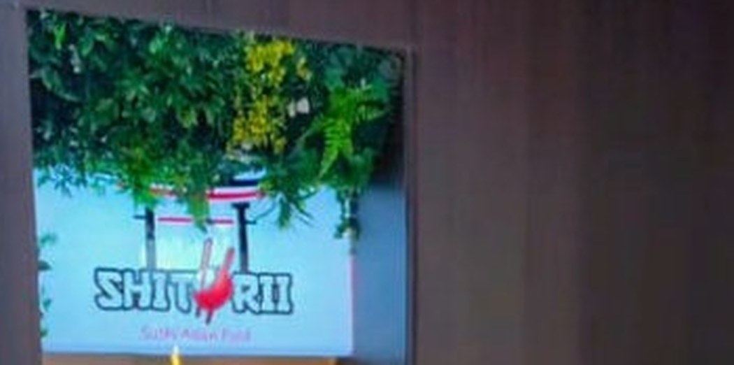 Letrero de neón de Shitorii entre plantas, junto a una pantalla azul