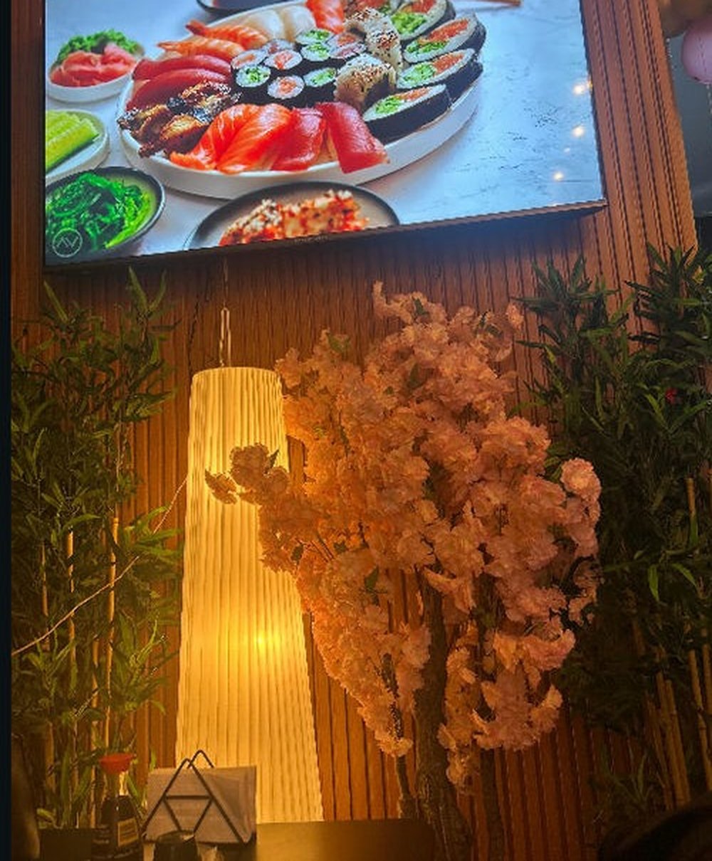 Interior de Shitorii Sushi: pantalla con una bandeja de sushi, lámpara de papel encendida, ramas de sakura y plantas